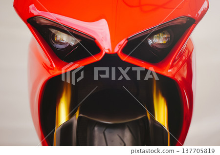 A tight macro shot of the aggressive front fairing and dual headlights of a modern red sportbike, highlighting its sleek design and golden fork tubes. A tight macro shot of the aggressive front fairing and dual headlights of a modern red sportbike, highlighting its sleek design and golden fork tubes. 137705819