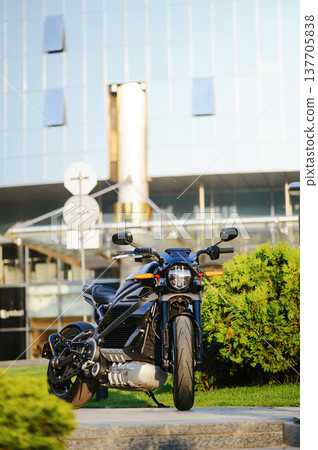 A vertical full-shot of a sleek black electric motorcycle parked on a sidewalk, featuring a glass office building and green bushes in the background. A vertical full-shot of a sleek black electric motorcycle parked on a sidewalk, featuring a glass office building and green bushes in the background. 137705838