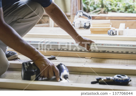 A carpenter's hands measuring the dimensions of lumber with a tape measure at a wooden house construction site. A carpenter's hands measuring the dimensions of lumber with a tape measure at a wooden house construction site. 137706044