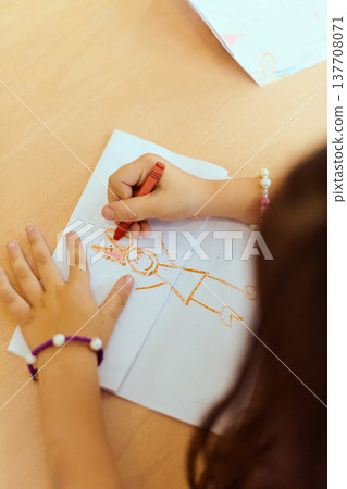 Child drawing with red crayon on paper at a classroom table, hands and simple stick figure artwork 137708071