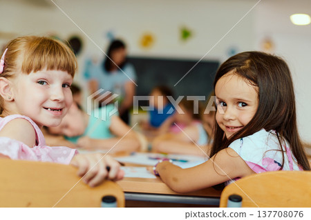 Two smiling girls sitting at a classroom table with classmates in the background 137708076