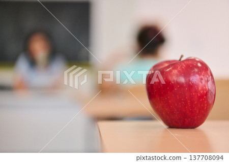 Red apple on a school desk in a classroom with blurred students and teacher in the background 137708094