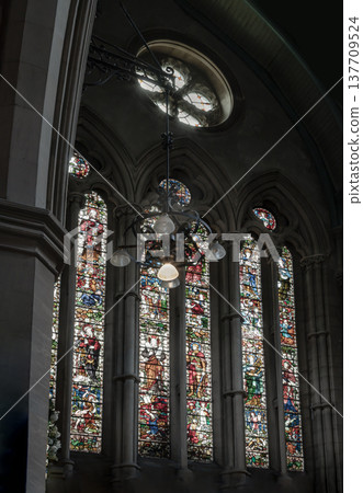 Sunlight filters through the vibrant stained-glass triptych of St. Mary Abbots, painting the ancient stone in a kaleidoscope of color. Above the sacred space, an ornate chandelier hangs from the Sunlight filters through the vibrant stained-glass triptych of St. Mary Abbots, painting the ancient stone in a kaleidoscope of color. Above the sacred space, an ornate chandelier hangs from the 137709524