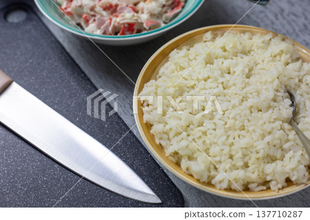 Large bowl of cooked rice with spoon surrounded by sushi ingredients, cutting board nearby. Another dish with crab sticks mixed with mayonnaise. Preparation for Asian homemade meal Large bowl of cooked rice with spoon surrounded by sushi ingredients, cutting board nearby. Another dish with crab sticks mixed with mayonnaise. Preparation for Asian homemade meal 137710287
