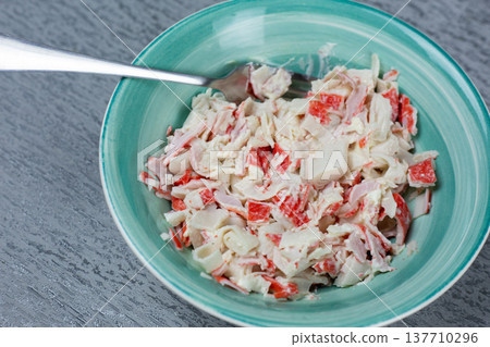 Bowl filled with crab salad featuring imitation crab meat, mayonnaise, and diced vegetables, placed on a textured gray surface with a fork positioned on the edge of the bowl 137710296