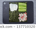 Ingredients for sushi preparation displayed on a black cutting board, including nori, cucumber slices, imitation crab meat, and cream cheese, arranged in distinct sections for easy assembly 137710320