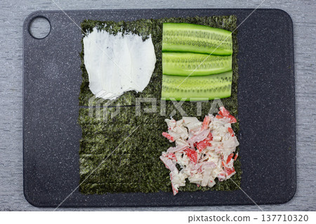 Ingredients for sushi preparation displayed on a black cutting board, including nori, cucumber slices, imitation crab meat, and cream cheese, arranged in distinct sections for easy assembly 137710320