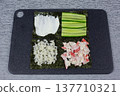 Ingredients for sushi preparation are arranged on a black cutting board, including sliced cucumber, crab meat, rice, and nori seaweed, each placed in distinct sections for easy access 137710321