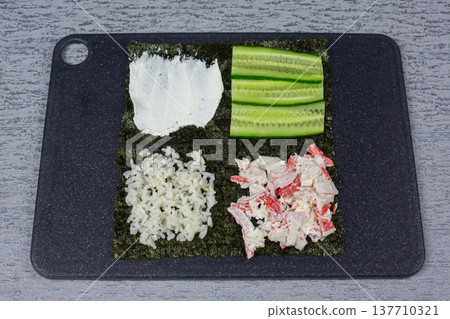 Ingredients for sushi preparation are arranged on a black cutting board, including sliced cucumber, crab meat, rice, and nori seaweed, each placed in distinct sections for easy access Ingredients for sushi preparation are arranged on a black cutting board, including sliced cucumber, crab meat, rice, and nori seaweed, each placed in distinct sections for easy access 137710321