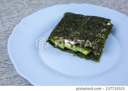 Seaweed wrap containing cucumber and cream cheese presented on a light blue plate, with a textured gray surface providing a contrasting backdrop for the dish 137710325