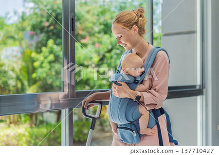 Woman vacuuming a room while carrying her baby in an ergonomic carrier. Modern motherhood, multitasking parent, home chores with infant, balance of childcare and housework concept. 137710427