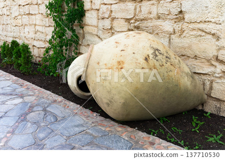 A large, brown, ceramic vase is sitting on the ground next to some plants A large, brown, ceramic vase is sitting on the ground next to some plants 137710530