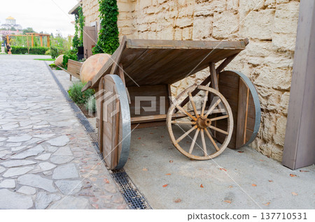 A wooden cart with a wheel is leaning against a stone wall 137710531