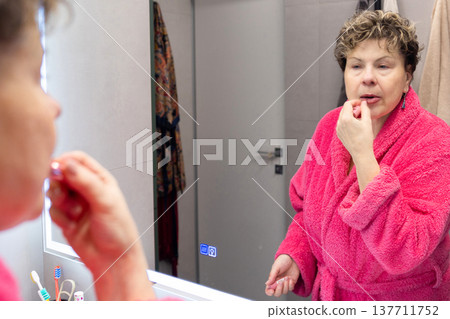 Mature woman applying lip balm in bathroom mirror at home Mature woman applying lip balm in bathroom mirror at home 137711752