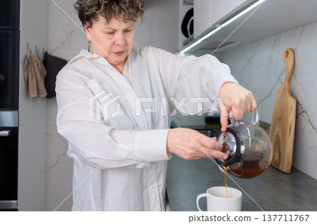 Senior woman pouring tea into cup in modern kitchen Senior woman pouring tea into cup in modern kitchen 137711767
