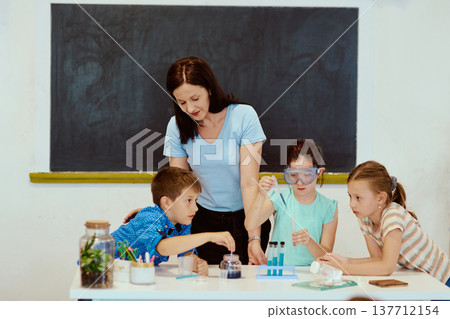 Teacher guides children through a hands-on science experiment in classroom with test tubes and goggles Teacher guides children through a hands-on science experiment in classroom with test tubes and goggles 137712154