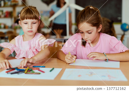 Two schoolgirls drawing with crayons at a classroom table focused on art and learning activities Two schoolgirls drawing with crayons at a classroom table focused on art and learning activities 137712162