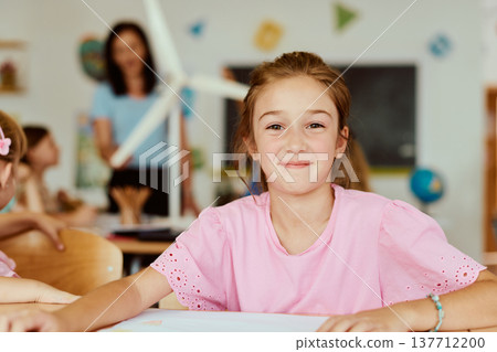 Smiling schoolgirl sitting at desk in bright classroom during creative lesson with teacher in background Smiling schoolgirl sitting at desk in bright classroom during creative lesson with teacher in background 137712200