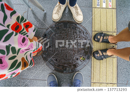 Osaka, Japan - Sep 22 2024, close-up view of metal manhole with patterns depicting a bridge over a river, buildings and a pagoda, surrounded by legs of four people, at Dotonbori district, Osaka, Japan Osaka, Japan - Sep 22 2024, close-up view of metal manhole with patterns depicting a bridge over a river, buildings and a pagoda, surrounded by legs of four people, at Dotonbori district, Osaka, Japan 137713293