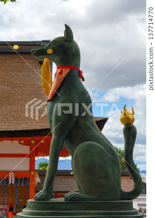 Kyoto, Japan - Sep 23 2024, Close-up view of the stone statue of kitsune, a Japanese fox holding a golden object in its mouth, with red facade of Fushimi-Inari Temple in background, Kyoto, Japan Kyoto, Japan - Sep 23 2024, Close-up view of the stone statue of kitsune, a Japanese fox holding a golden object in its mouth, with red facade of Fushimi-Inari Temple in background, Kyoto, Japan 137714770
