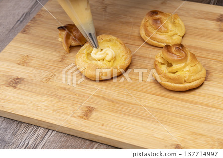 特寫鏡頭展現了廚房裡製作泡芙的手:將奶油餡擠入泡芙麵團中,並撒上糖粉。 特寫鏡頭展現了廚房裡製作泡芙的手:將奶油餡擠入泡芙麵團中,並撒上糖粉。 137714997