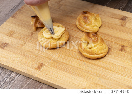 特寫鏡頭展現了廚房裡製作泡芙的手:將奶油餡擠入泡芙麵團中,並撒上糖粉。 特寫鏡頭展現了廚房裡製作泡芙的手:將奶油餡擠入泡芙麵團中,並撒上糖粉。 137714998