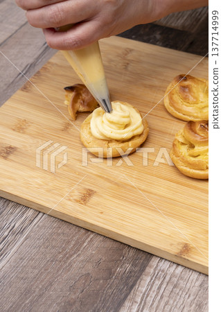 特寫鏡頭展現了廚房裡製作泡芙的手:將奶油餡擠入泡芙麵團中,並撒上糖粉。 特寫鏡頭展現了廚房裡製作泡芙的手:將奶油餡擠入泡芙麵團中,並撒上糖粉。 137714999