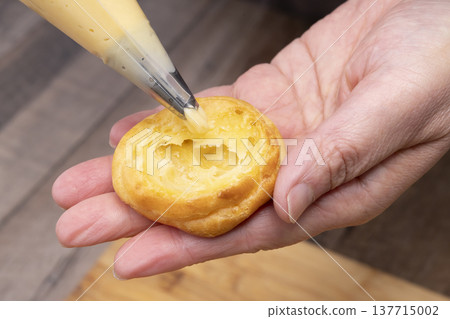 特寫鏡頭展現了廚房裡製作泡芙的手:將奶油餡擠入泡芙麵團中,並撒上糖粉。 特寫鏡頭展現了廚房裡製作泡芙的手:將奶油餡擠入泡芙麵團中,並撒上糖粉。 137715002