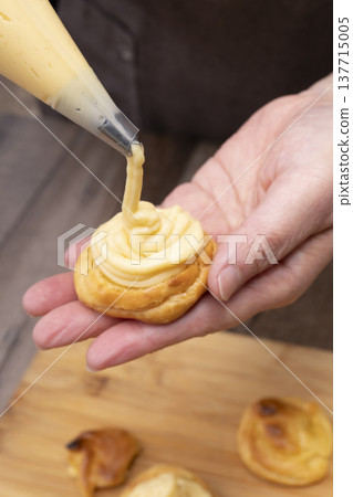 特寫鏡頭展現了廚房裡製作泡芙的手:將奶油餡擠入泡芙麵團中,並撒上糖粉。 特寫鏡頭展現了廚房裡製作泡芙的手:將奶油餡擠入泡芙麵團中,並撒上糖粉。 137715005