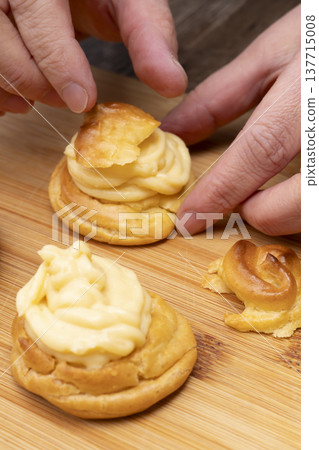 特寫鏡頭展現了廚房裡製作泡芙的手:將奶油餡擠入泡芙麵團中,並撒上糖粉。 特寫鏡頭展現了廚房裡製作泡芙的手:將奶油餡擠入泡芙麵團中,並撒上糖粉。 137715008