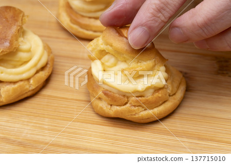 特寫鏡頭展現了廚房裡製作泡芙的手:將奶油餡擠入泡芙麵團中,並撒上糖粉。 特寫鏡頭展現了廚房裡製作泡芙的手:將奶油餡擠入泡芙麵團中,並撒上糖粉。 137715010