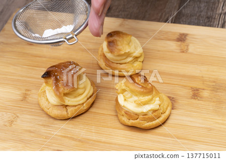 特寫鏡頭展現了廚房裡製作泡芙的手:將奶油餡擠入泡芙麵團中,並撒上糖粉。 特寫鏡頭展現了廚房裡製作泡芙的手:將奶油餡擠入泡芙麵團中,並撒上糖粉。 137715011