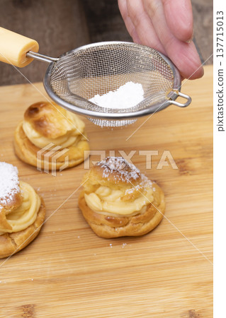 特寫鏡頭展現了廚房裡製作泡芙的手:將奶油餡擠入泡芙麵團中,並撒上糖粉。 特寫鏡頭展現了廚房裡製作泡芙的手:將奶油餡擠入泡芙麵團中,並撒上糖粉。 137715013