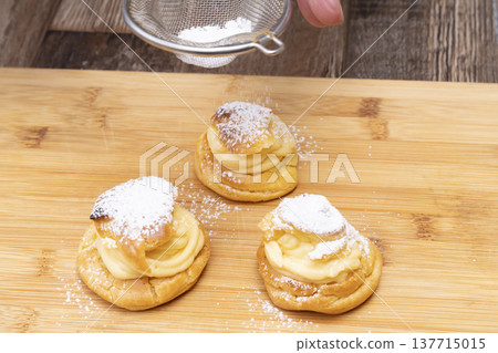 特寫鏡頭展現了廚房裡製作泡芙的手:將奶油餡擠入泡芙麵團中,並撒上糖粉。 特寫鏡頭展現了廚房裡製作泡芙的手:將奶油餡擠入泡芙麵團中,並撒上糖粉。 137715015