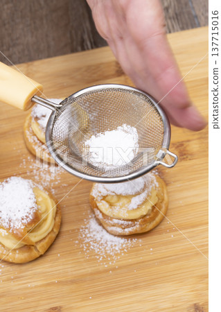 特寫鏡頭展現了廚房裡製作泡芙的手:將奶油餡擠入泡芙麵團中,並撒上糖粉。 特寫鏡頭展現了廚房裡製作泡芙的手:將奶油餡擠入泡芙麵團中,並撒上糖粉。 137715016