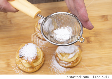 特寫鏡頭展現了廚房裡製作泡芙的手:將奶油餡擠入泡芙麵團中,並撒上糖粉。 特寫鏡頭展現了廚房裡製作泡芙的手:將奶油餡擠入泡芙麵團中,並撒上糖粉。 137715017