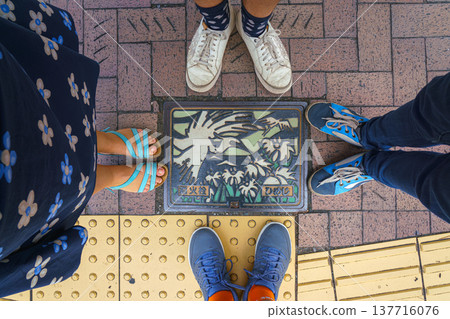 Himeji, Japan - Sep 24 2024, A close-up view of a manhole with colorful picture of stylized flying white egrets over a river and a field of reeds, surrounded by feet of a family of four, Himeji, Japan Himeji, Japan - Sep 24 2024, A close-up view of a manhole with colorful picture of stylized flying white egrets over a river and a field of reeds, surrounded by feet of a family of four, Himeji, Japan 137716076