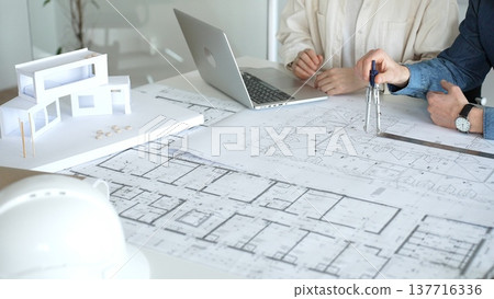 Architects collaborating on architectural blueprints and building plans, designing a new house project with a model, laptop, and safety helmet on a desk in an office 137716336