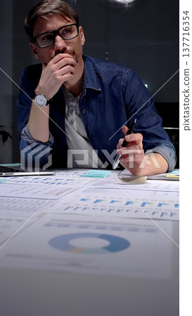 Professional man in glasses thinking deeply while analyzing financial charts and data on an office desk, working late night in a dark setting, feeling focused and determined 137716354