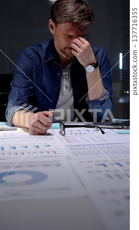 Young businessman with glasses sits at a dark office desk late at night, overwhelmed by documents and a tablet, clutching his head from stress, burnout and exhaustion Young businessman with glasses sits at a dark office desk late at night, overwhelmed by documents and a tablet, clutching his head from stress, burnout and exhaustion 137716355