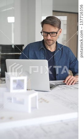 Male architect thinking while looking at a scale model of a modern house, developing architectural plans and blueprints for a new residential project in an office setting 137716356