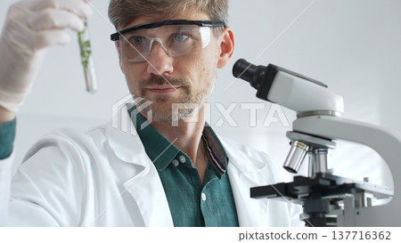 Adult male scientist in mask and gloves holding test tube with green plant sample, conducting laboratory research with a microscope. Medicine, healthcare and science concept Adult male scientist in mask and gloves holding test tube with green plant sample, conducting laboratory research with a microscope. Medicine, healthcare and science concept 137716362