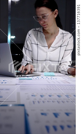 Businesswoman working late night in a modern office, analyzing financial data on a digital tablet and reviewing paperwork, demonstrating dedication and overtime productivity 137716391