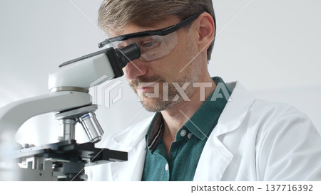 Senior man scientist wearing protective glasses and lab coat analyzing samples with a microscope in laboratory setting. Medicine, health care and science concept Senior man scientist wearing protective glasses and lab coat analyzing samples with a microscope in laboratory setting. Medicine, health care and science concept 137716392
