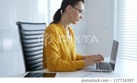 Professional businesswoman wearing yellow shirt and eyeglasses working intently, typing on laptop in bright office space with documents and tablet surrounding workspace. Business people concept 137716397
