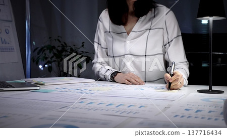 Businesswoman working late under a desk lamp, reviewing charts and financial documents with focused concentration, preparing reports and planning strategy for company growth 137716434