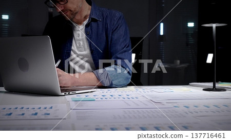 Man working late in a dim office, studying financial reports and charts across his desk while using a laptop, focused on deadlines, strategy and business growth Man working late in a dim office, studying financial reports and charts across his desk while using a laptop, focused on deadlines, strategy and business growth 137716461