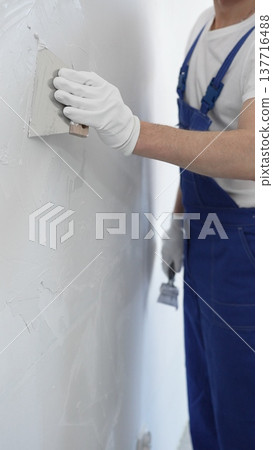 Unknown man construction worker wearing gloves and blue overalls is using a putty knife to apply plaster on a white wall, performing home renovation and improvement Unknown man construction worker wearing gloves and blue overalls is using a putty knife to apply plaster on a white wall, performing home renovation and improvement 137716488