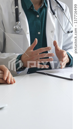 Doctor man wearing medical coat over green shirt, and stethoscope is gesturing with hands while explaining diagnosis to patient during medical consultation in clinic office. Medicine and health care 137716499