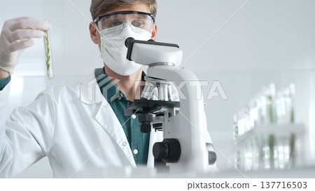 Masked adult man researcher examining green botanical samples in test tube in a sterile laboratory during biological analysis. Medicine, healthcare and science concept 137716503